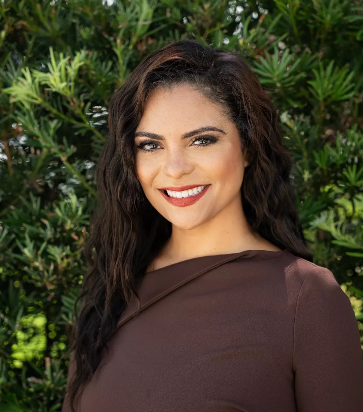 A woman with long, wavy dark hair and a brown dress smiles at the camera, standing outdoors in front of green leafy bushes.