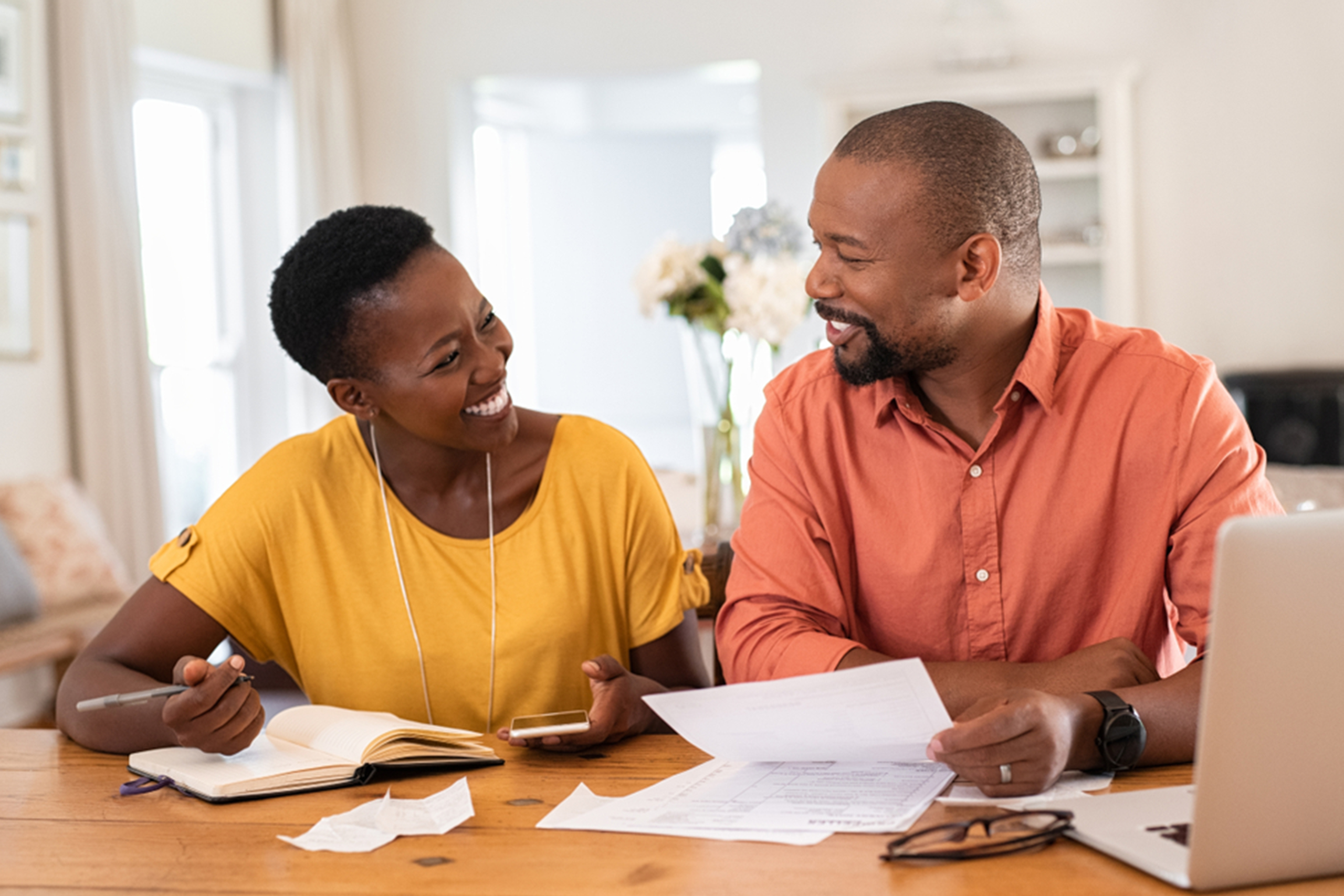 A smiling couple sits at a table, discussing paperwork. The woman, in a yellow shirt, holds a pen and notebook, while the man, in an orange shirt, holds documents. A laptop and scattered papers are on the table, suggesting a home office setting.