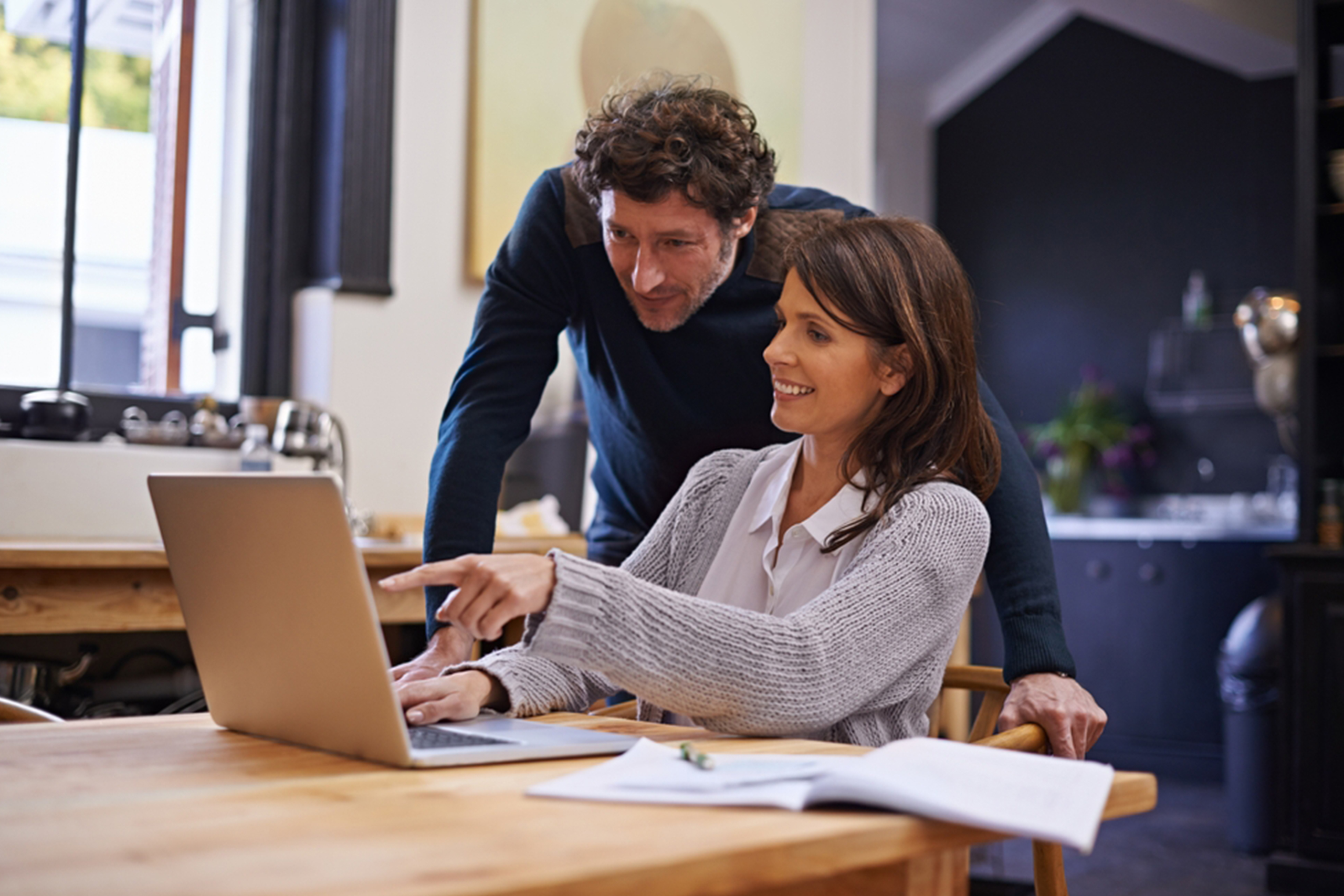 A woman sitting at a table in a kitchen points at a laptop screen, smiling. A man stands next to her, leaning over her shoulder and also looking at the laptop screen. They both appear engaged and interested in the content.