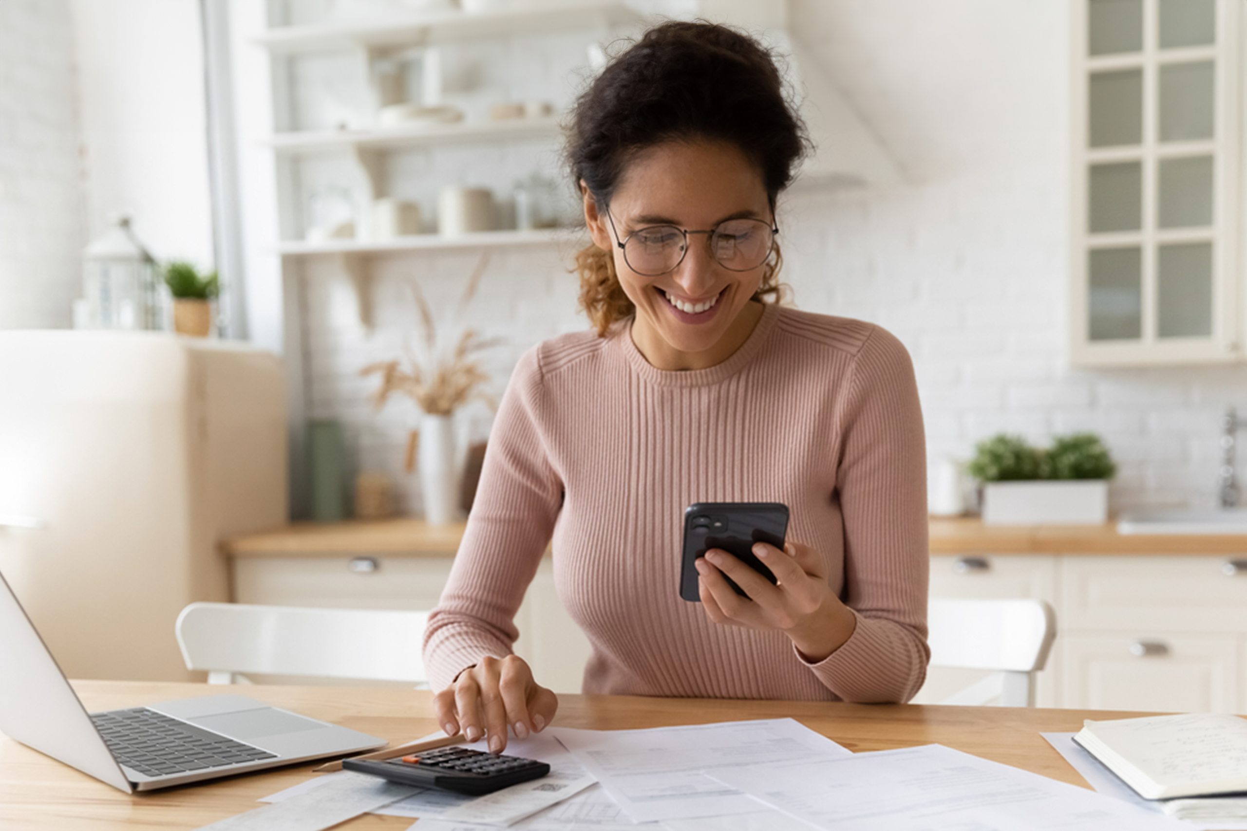 A woman wearing glasses and a pink sweater sits at a table using a smartphone. A laptop, calculator, and papers are on the table. She appears happy and the setting is a bright kitchen.