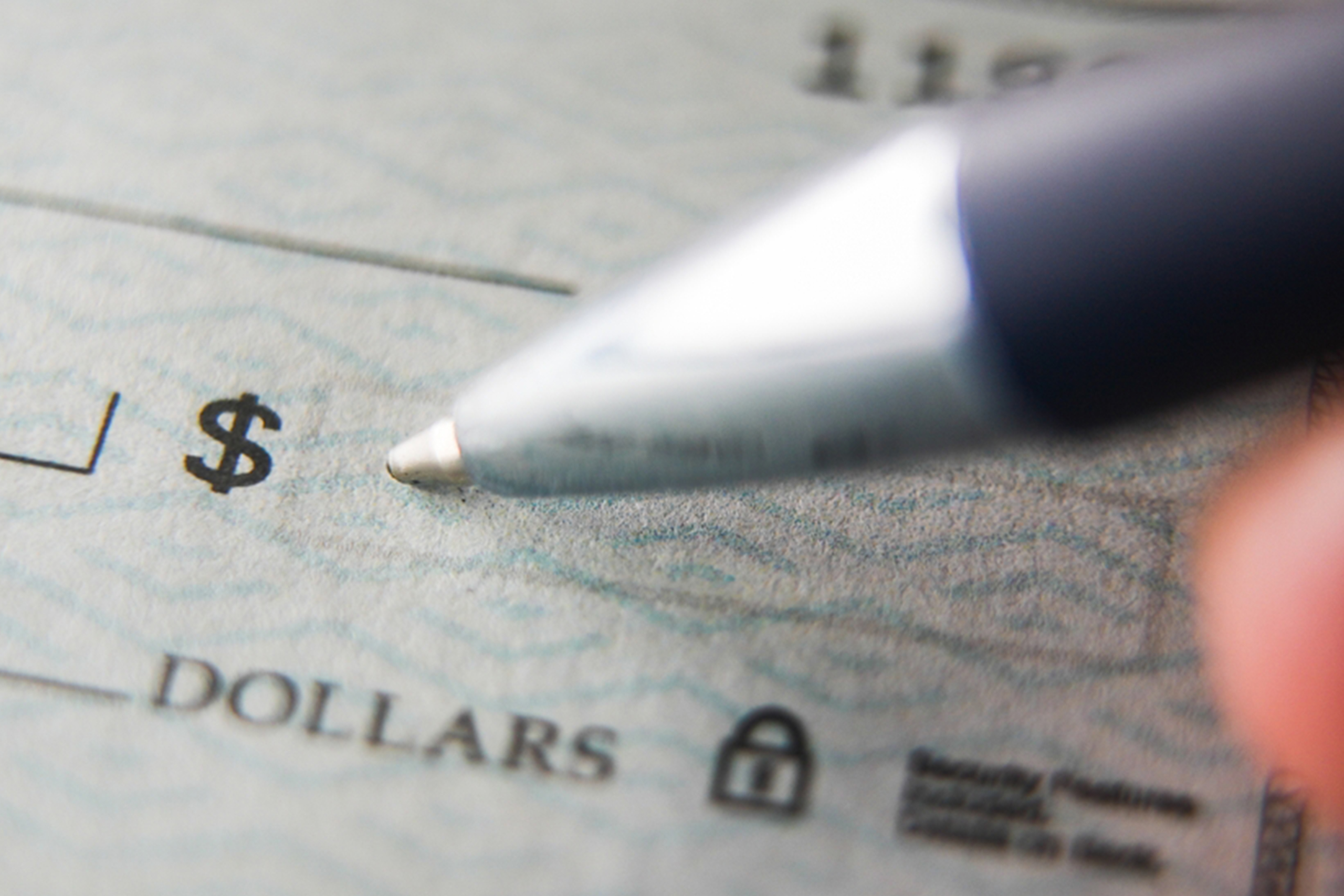 Close-up of a pen poised above a blank check. The check features a dollar sign and a security padlock symbol, indicating a secure document. The background shows a textured pattern typical of check paper.