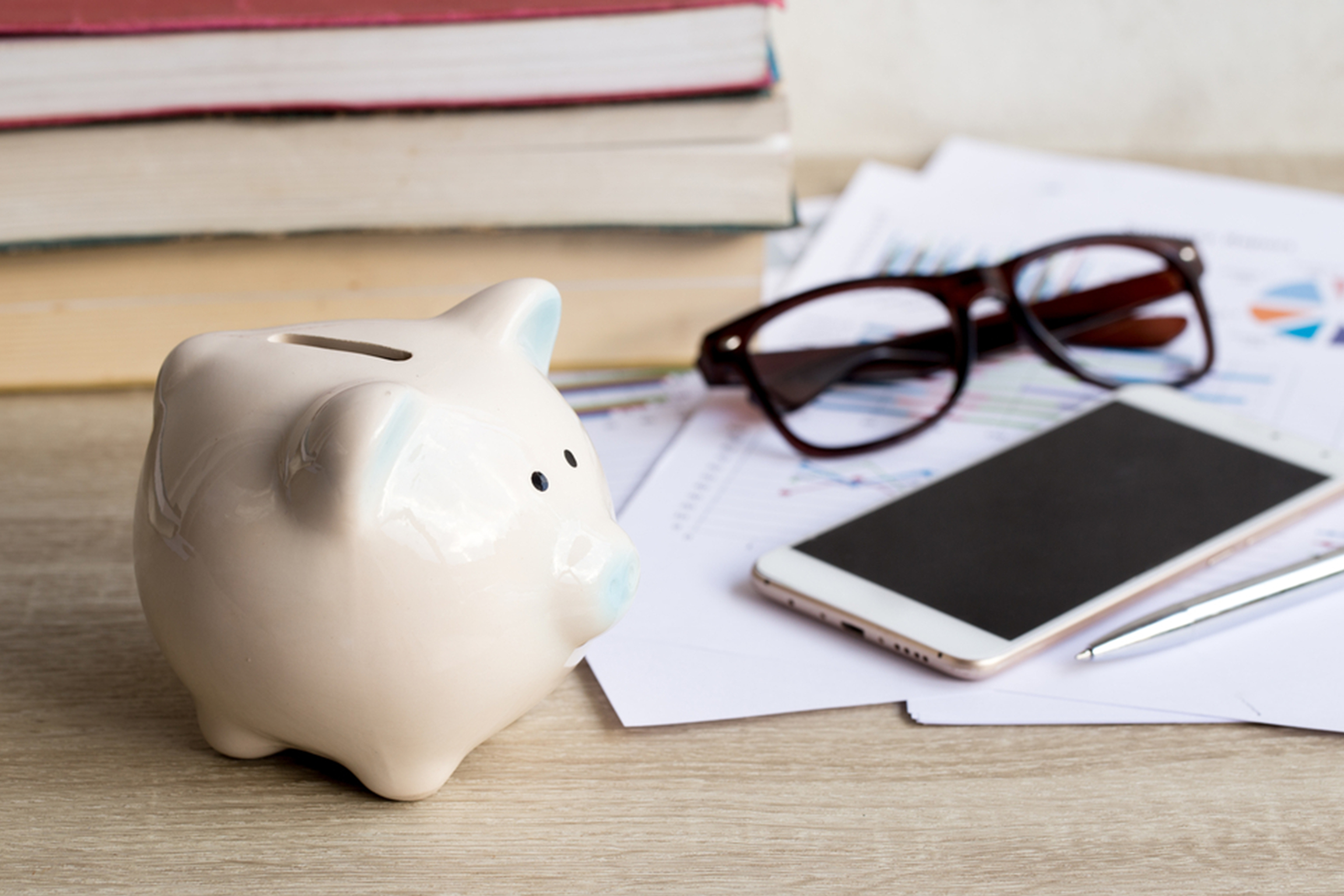 A white piggy bank sits on a desk beside a stack of books and papers. Nearby are a pair of glasses, a smartphone, and a pen. The scene suggests a focus on saving and financial planning.