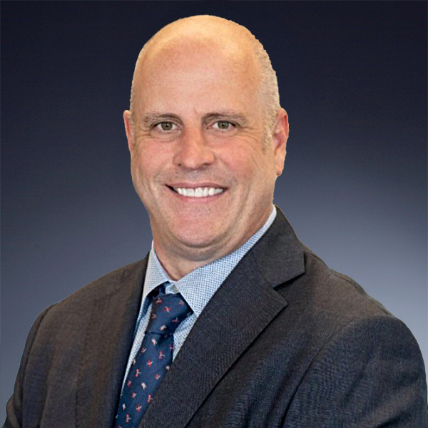 A smiling man in a suit and tie stands against a gradient dark background. He is bald and wearing a light blue dress shirt with a patterned tie.