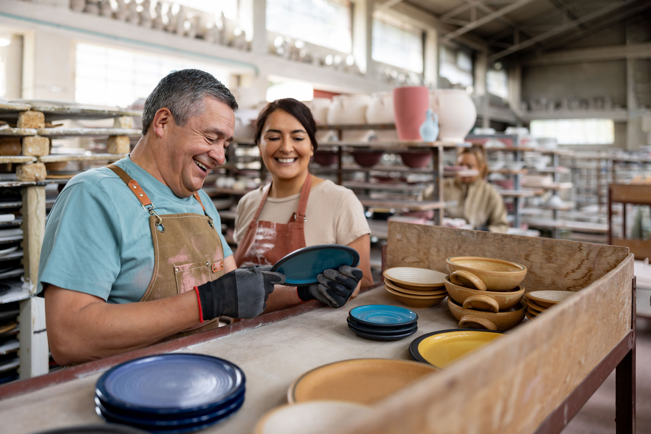Two people wearing aprons smile and examine ceramic plates in a pottery studio, surrounded by shelves filled with various pottery items. The atmosphere is bright and cheerful.