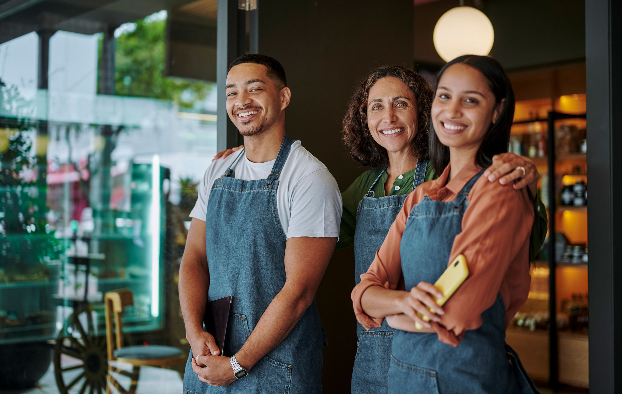 Three people wearing blue aprons stand together at the entrance of a shop, smiling at the camera. One person holds a yellow smartphone. Shelves and outdoor scenery are visible in the background.
