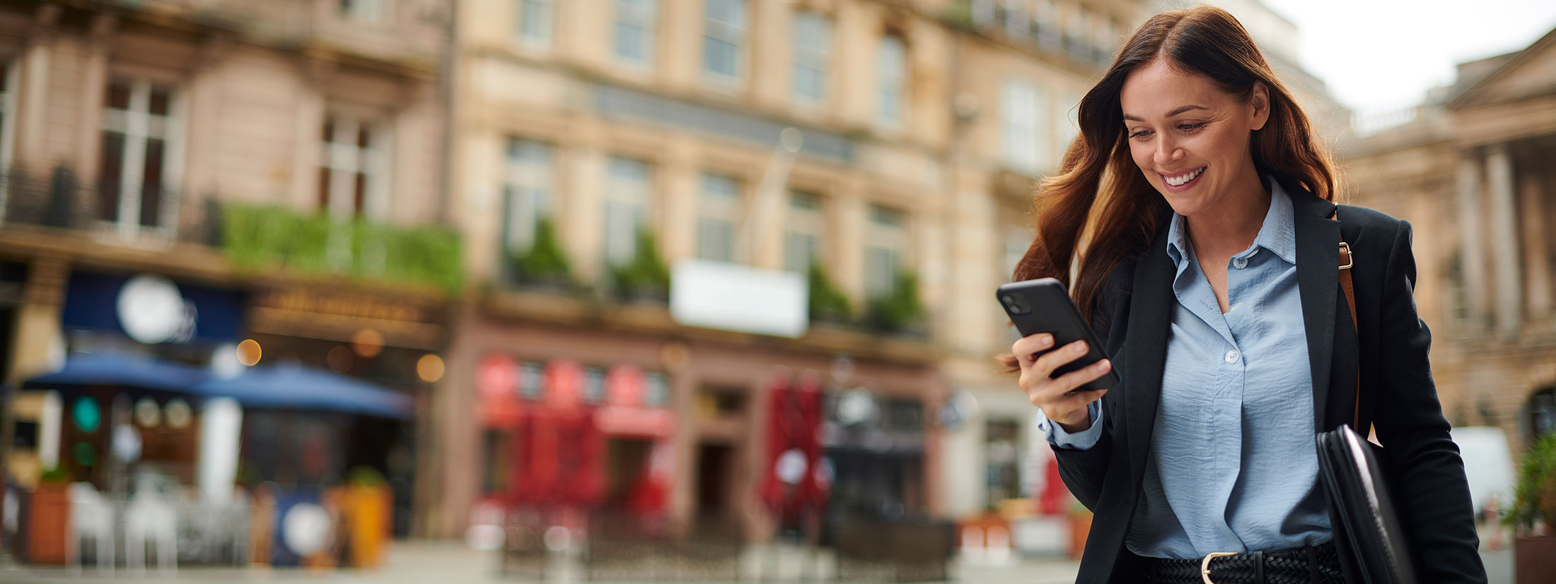 A smiling woman in business attire walks outside in a city, holding a folder and looking at her smartphone; blurred buildings and shops are visible in the background.