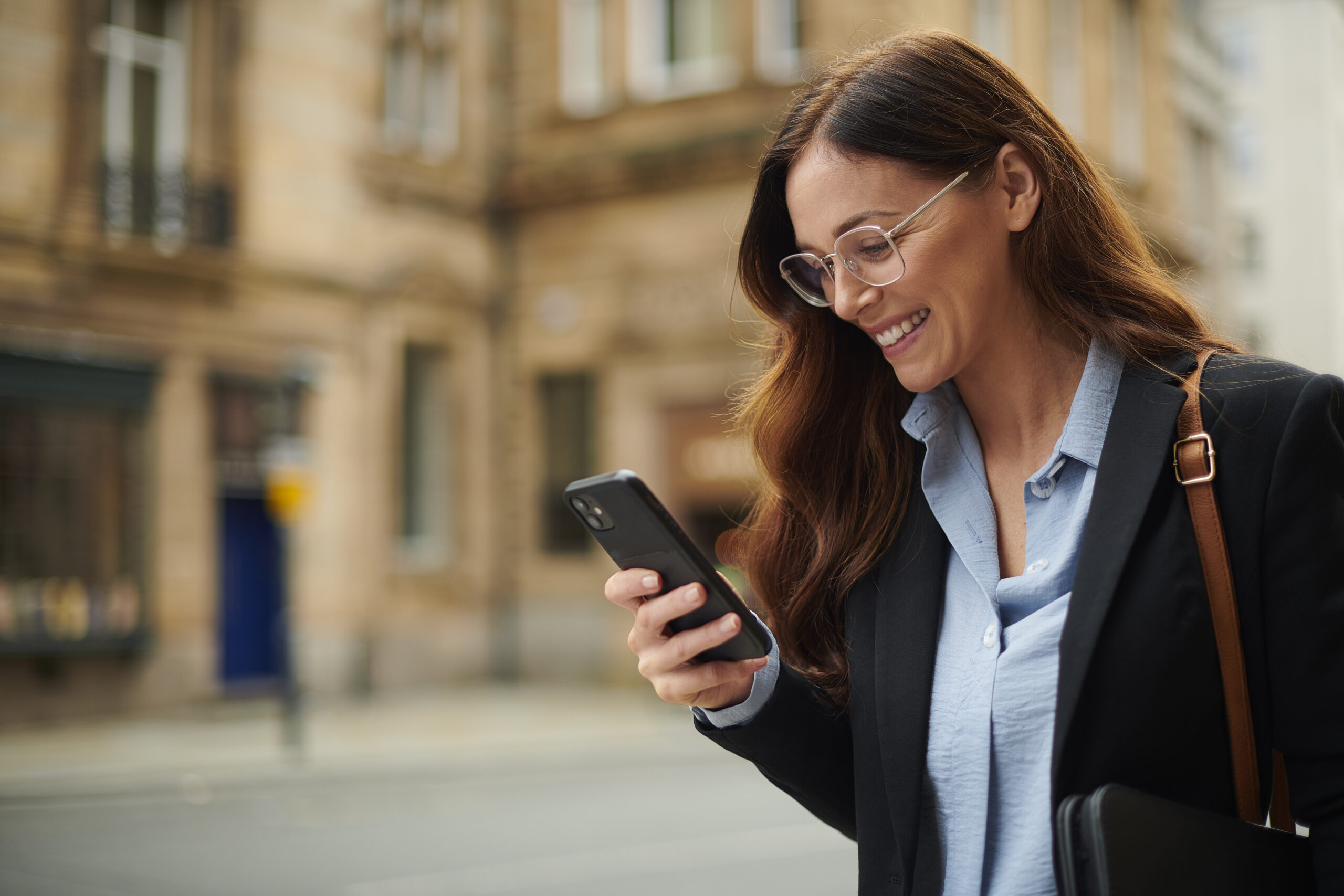 A woman with long brown hair and glasses smiles while looking at her smartphone on a city street. She is wearing a blue shirt, black blazer, and carrying a brown-strap bag. The background is slightly blurred.