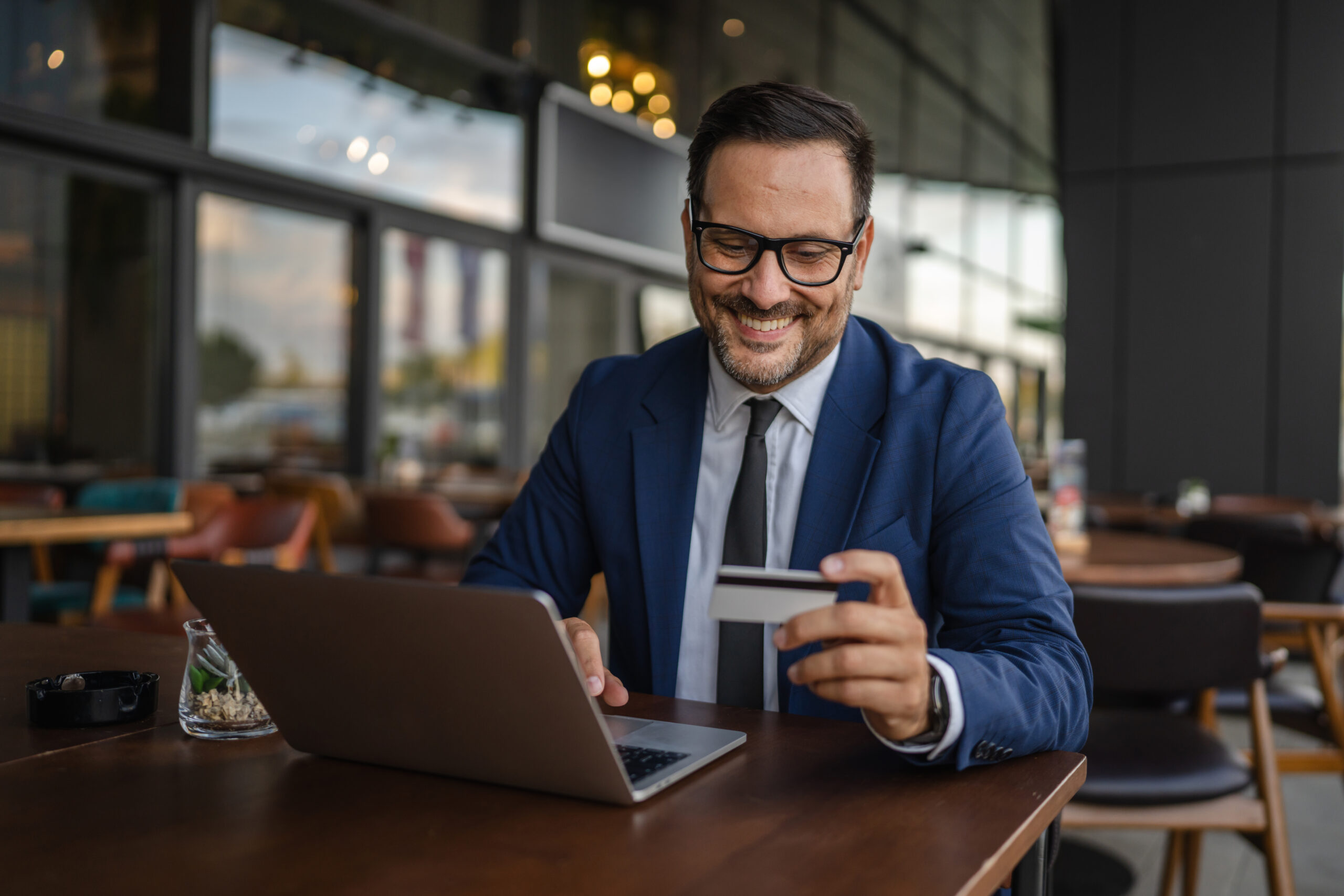 A smiling man in a blue suit and glasses sits at a table in a modern cafe, holding a credit card and using a laptop, suggesting he is shopping or banking online.