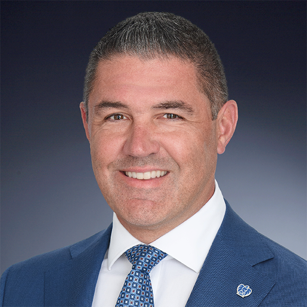 Smiling man in a blue suit and patterned tie poses against a dark gradient background. He has short dark hair and is wearing a pin on his lapel.