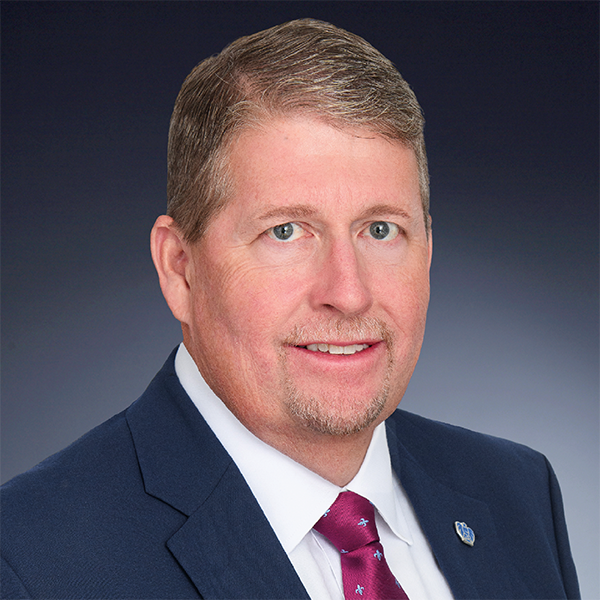 A man with short light brown hair and a goatee, wearing a navy suit, white shirt, and maroon tie, poses against a gradient dark blue background.