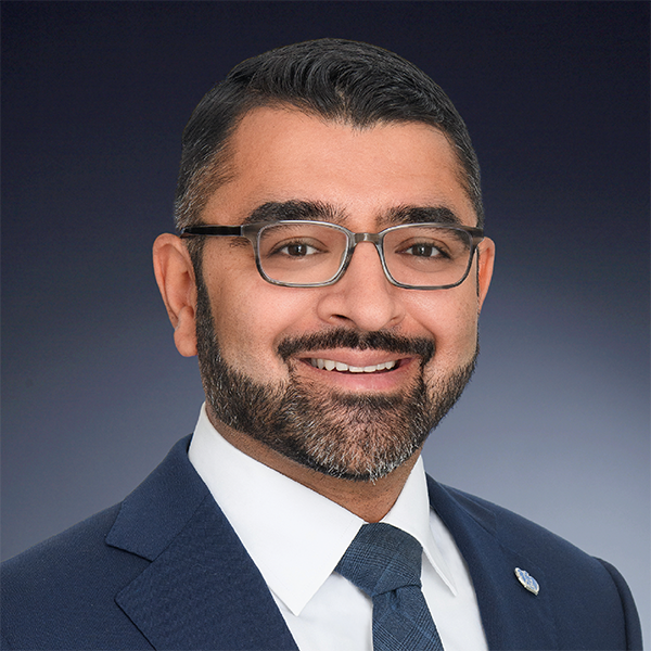 A man with short dark hair, glasses, and a beard, wearing a navy suit, white shirt, and blue tie, smiling in a professional studio portrait with a dark gradient background.