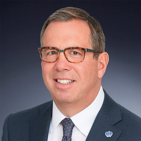 A man wearing glasses, a blue suit, white shirt, and polka dot tie, smiling in front of a dark gradient background. A blue and white lapel pin is visible on his jacket.